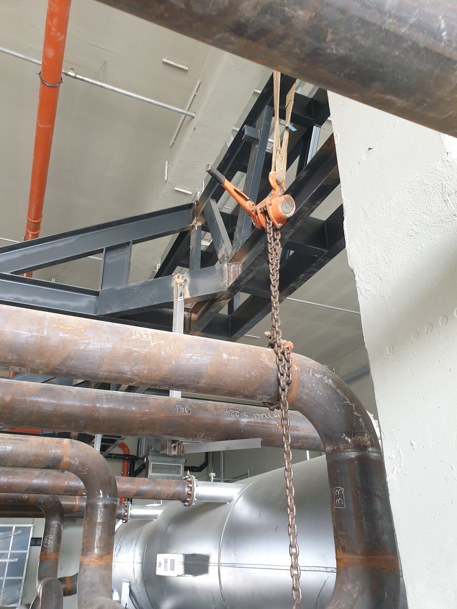 Palan cu lanț și chingi montate lângă conducte și structura metalică de susținere.